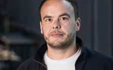 Professional portrait of a man in casual business attire standing in a modern office environment with soft lighting and a blurred background.