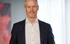 Porträtt av en man i mörk kavaj och vit skjorta, stående inomhus.