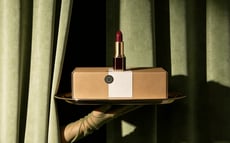 A serving hand holding a tray with a red lipstick on a box in front of green curtains.