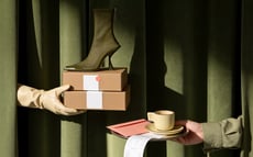 Olive-green heeled ankle boot on boxes, hands emerging from green curtains with coffee cup and receipt.