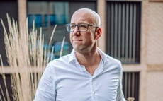 Kale man met een bril en een wit overhemd die buiten staat en bedachtzaam opzij kijkt, met een modern gebouw op de achtergrond