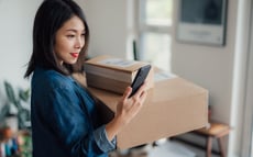 Eine Frau, die zwei Kartonschachteln in ihrem linken Arm trägt, schaut auf ihr Smartphone, während sie sich drinnen befindet. Sie trägt eine blaue Jacke und lächelt leicht. Im Hintergrund ist ein sanft beleuchteter Raum mit einem verschwommenen Schreibtisch und einem Fenster zu sehen.