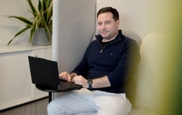 A person sits in a high-backed, cushioned office booth, using a black laptop placed on a small round table. They are wearing a dark navy zip-collar sweater, light-colored pants, and a smartwatch. A potted plant with long green leaves sits on a cabinet in the background, suggesting a modern, quiet workspace designed for focused work.