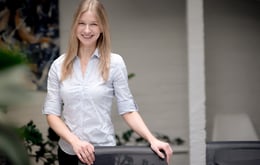 A smiling woman with long blonde hair stands indoors, wearing a light blue button-up shirt and resting her hands on the back of a chair in a bright office setting.