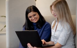 Two women sit together indoors, smiling and looking at a laptop—one wearing glasses and a blue sweater, the other with long blonde hair in a light shirt—suggesting a collaborative or friendly work moment.