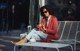 nainen istuu ulkona nauttien kahvista ja käyttäen puhelinta