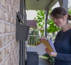 Eine Frau hält einen Brief in der Hand und schaut neugierig auf den Briefkasten.