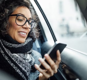 Smilende kvinne med briller sitter i en bil og bruker smarttelefonen sin til å håndtere fakturaer mens hun er på farten.