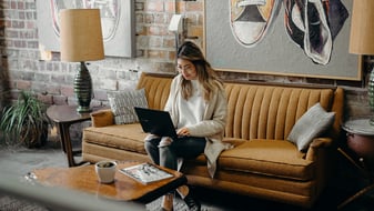 Eine junge Frau sitzt auf einem senffarbenen Sofa und schaut auf ihren Laptop