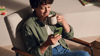 Person sitting relaxed in an armchair, looking at a smartphone and holding a mug.