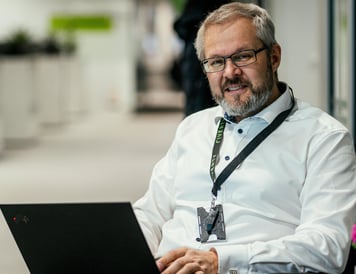 A professional in a white shirt working on a laptop in a modern office setting, smiling at the camera.