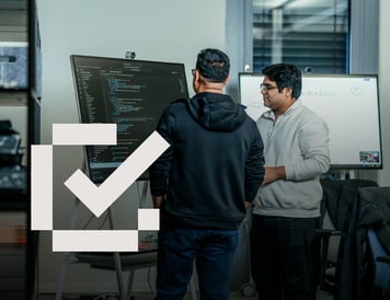 Checklist item marked as complete. Two colleagues collaborating in an office, discussing software development work displayed on a large digital screen. The image captures teamwork, knowledge sharing, and problem-solving in a technical environment.