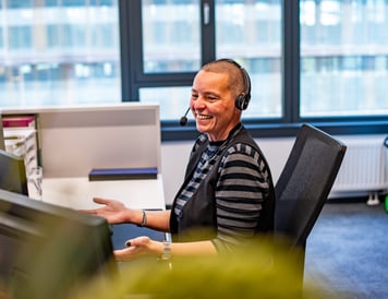 A smiling customer service representative wearing a headset, engaged in a conversation at their desk