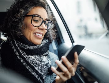 Smiling woman with glasses sitting in a car, using her smartphone to manage invoices on the go.