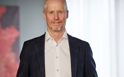 Porträtt av en man i mörk kavaj och vit skjorta, stående inomhus.