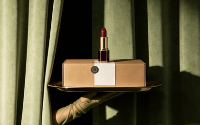 Eine servierende Hand hält ein Tablett mit einem roten Lippenstift auf einer Box vor grünen Vorhängen