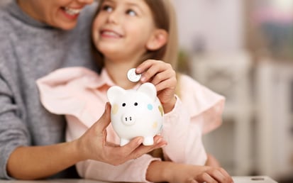 A little girl sits on her mom's lap while mom holds a piggy bank in her hand and the little girl puts one euro in the piggy bank
