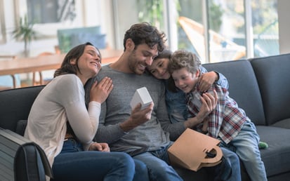 Eine Mama und ein Papa sitzen gemeinsam mit ihrem Sohn und ihrer Tochter auf dem Sofa übergeben Geschenke und umarmen sich