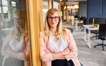 A blonde woman with glasses, wearing a light pink blazer and white top, leans with folded arms against a wooden doorframe, with her reflection visible on a adjacent glass panel in a modern office space.