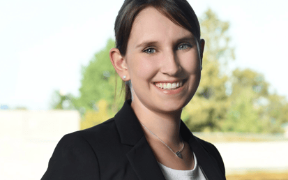 At the center of the image, a person is facing the camera and smiling warmly. The person is wearing a black blazer over a light-colored top. Their hair is brown, pulled back, with a slightly off-center part. Visible accessories include small stud earrings and a delicate necklace with a small leaf- or teardrop-shaped pendant. The background is blurred and shows trees and greenery outdoors, with a light horizontal area in the lower part of the image. No text, logos, or brand markings are visible. The overall impression is that of a professional portrait taken outside.