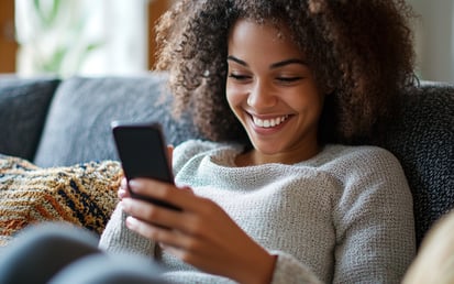 Een vrouw zit op de bank lachend en kijkt op haar telefoon