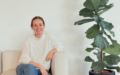 Eine lächelnde Person sitzt auf einem hellen Sofa, trägt einen weißen Pullover und Jeans, und neben ihr steht eine große, grüne Pflanze in einem Topf vor einer schlichten Wand.