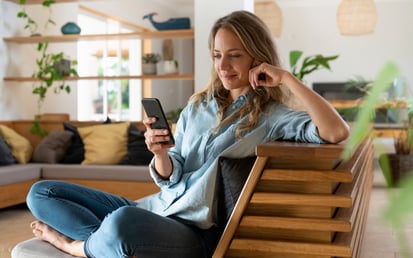 Eine Dame sitzt lächelnd auf der Couch und schaut auf ihr Telefon
