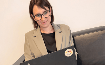 Eine Person mit dunklem, etwa schulterlangem Haar und dunkler Brille sitzt auf einem schwarzen Ledersofa vor einer hellen, einfarbigen Wand. Die Person trägt einen beigefarbenen Blazer über einem schwarzen Oberteil und blickt nach unten auf einen geöffneten schwarzen Laptop. Auf dem Laptop ist links das „ThinkPad“-Logo zu sehen. Oben rechts befindet sich ein runder Aufkleber mit einem mehrfarbigen, abstrakten Symbol. Die Aufnahme ist hell und in warmen, neutralen Farbtönen gehalten.