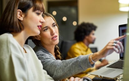Zwei junge Frauen sitzen vor einem PC-Bildschirm. Die eine zeigt auf etwas. 
