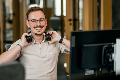 Ein junger Mann mit Brille und einem lächelnden Gesichtsausdruck sitzt an einem Schreibtisch in einem Büro. Er hält ein Paar Kopfhörer um seinen Hals und trägt ein hellbeiges Hemd. Am Handgelenk trägt er eine Smartwatch und am Unterarm ist ein Tattoo sichtbar. Vor ihm steht ein Computermonitor, und im Hintergrund sind Bürofenster, die für eine helle und freundliche Atmosphäre sorgen.