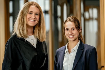 Zwei junge Frauen, die professionell gekleidet sind und in einem Bürokorridor stehen. Sie lächeln und blicken direkt in die Kamera. Die Frau auf der linken Seite trägt eine schwarze Jacke über einem weißen Oberteil und eine Halskette, während die Frau auf der rechten Seite ein dunkelblaues Jackett über einer weißen Bluse trägt. Der Hintergrund ist unscharf, mit großen Fenstern und Türen, die natürliches Licht hereinlassen.