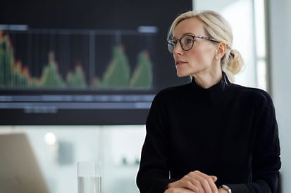 Businesswoman with blonde hair and glasses sitting at a table in front of a financial chart on a large screen