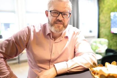 A man with short gray hair and glasses is sitting at a table in a modern office, wearing a light pink shirt. He is looking at the camera with a slight smile, with a coffee cup and smartphone in front of him.