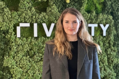 A woman with long blonde hair stands in front of a green plant-covered wall with a modern, partially visible white logo behind her. She is wearing a gray blazer over a dark top and looking directly at the camera with a neutral expression.