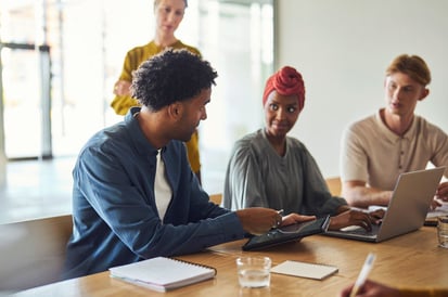 Vier Personen, an einem Konferenztisch sitzend, mit Laptops und Schreibmaterialien.