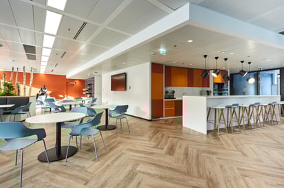Kantine mit einladender Atmosphäre, gekennzeichnet durch warme Farben an der Wand, vielfarbige Stühle und einen Holzboden.