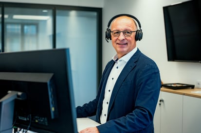 Mann mit Brille und Kopfhörern an einem Computerarbeitsplatz in einem modernen Büro mit mehreren Bildschirmen.