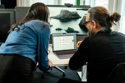 Zwei Personen arbeiten gemeinsam an einem Laptop in einem Büro; auf dem Bildschirm sind verschiedene grafische Symbole zu sehen.