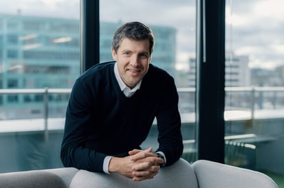 Man wearing a dark sweater over a white shirt, leaning forward and smiling while seated indoors with large windows overlooking city buildings.