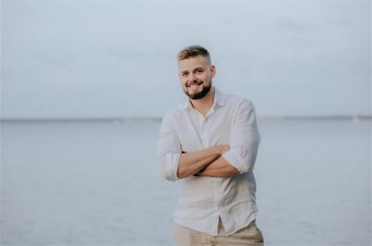 Man with short hair and beard wearing a light linen shirt and beige pants, standing with arms crossed and smiling by the water.