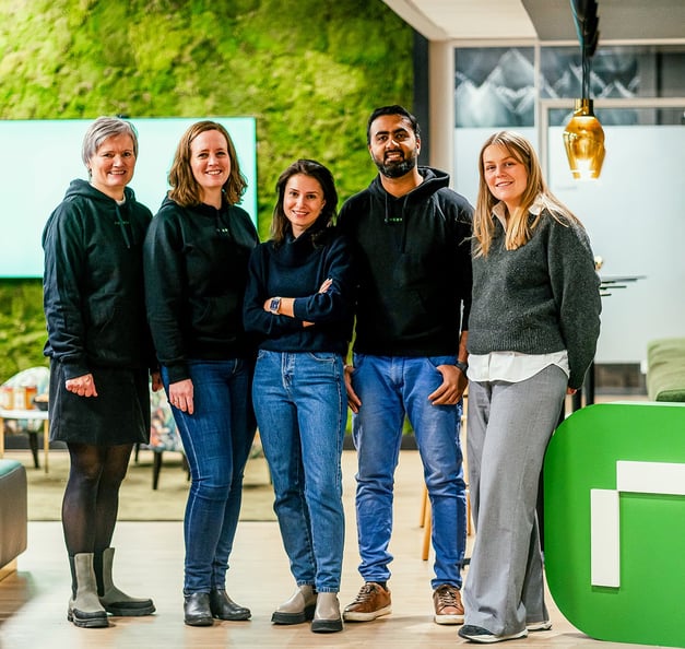 A group of five people standing together and smiling in a modern office with green wall décor and warm lighting. They appear relaxed and friendly, reflecting a collaborative and welcoming team environment.