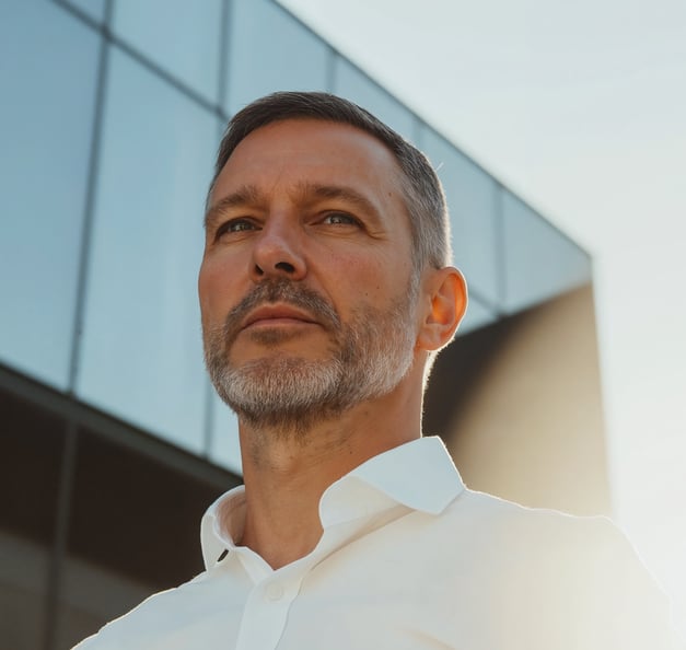 A businessman in a white shirt gazes ahead, focused, with a modern glass building in the background. Bright sunlight highlights his face.