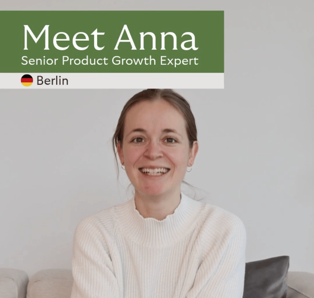 Eine Porträtaufnahme zeigt eine lächelnde Person, mittig im Bild, sitzend auf einem hellen Sofa vor einer schlichten, hellen Wand. Die Person trägt einen hellen, gestrickten Pullover; das Haar ist nach hinten gebunden. Rechts ist ein dunkles Sofakissen zu sehen. Oben im Bild liegt ein breiter, grüner Banner mit weißer Schrift: „Meet Anna“. Darunter steht „Senior Product Growth Expert“. Links daneben befindet sich ein kleines Deutschland-Flaggen-Icon, gefolgt von dem Wort „Berlin“. Das Bild wirkt wie eine Team- oder Profilvorstellung (Person wird als Expert:in vorgestellt).