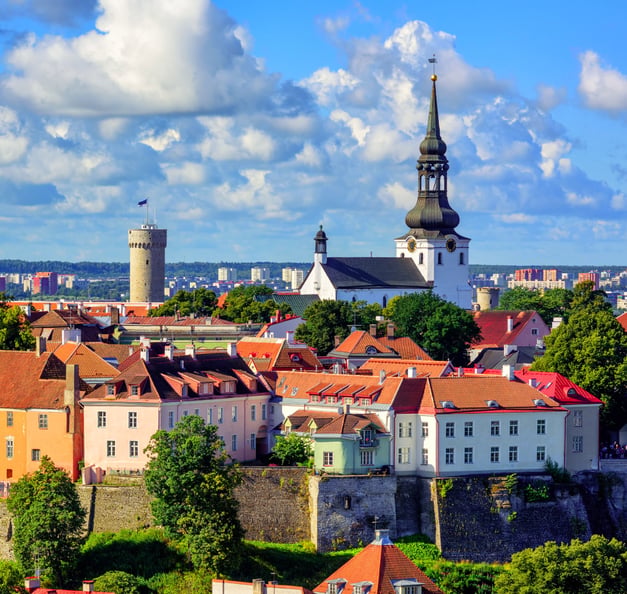 Eine farbenfrohe Stadtansicht von Tallinn mit historischen Gebäuden und roten Ziegeldächern, die dicht beieinander auf einer Anhöhe stehen. Im Zentrum ragt eine große Kirche mit einem hohen, spitzen Turm in den Himmel. Links daneben ist ein runder, mittelalterlicher Turm mit einer Flagge zu sehen. Im Hintergrund erstreckt sich eine moderne Stadtlandschaft unter einem blauen Himmel mit großen, weißen Wolken.