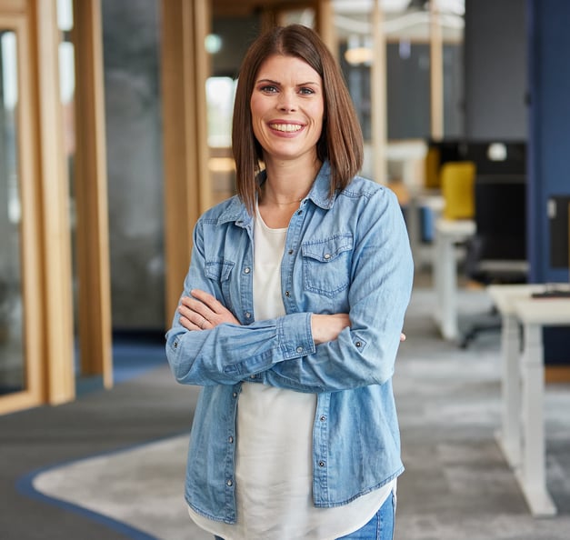 Eine Person mit schulterlangem braunem Haar steht in einem modernen Büro und lächelt mit verschränkten Armen. Sie trägt ein helles Jeanshemd über einem weißen Top; im Hintergrund sind Schreibtische und Glastüren zu sehen.