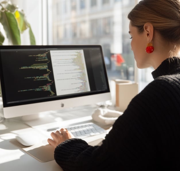 Frau arbeitet an einem iMac mit Code-Editor auf dem Bildschirm, im Hintergrund Fensterfront