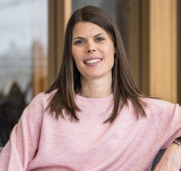 The image shows Carina seated in a relaxed posture, likely on a chair or armchair. The person has long, straight, dark brown hair worn open, falling onto their shoulders. Their facial expression is friendly and open: they are smiling naturally into the camera, conveying approachability and ease. The person is wearing a pink knitted sweater with a round neckline and long sleeves. A bracelet or watch is partially visible at the wrist. Their body posture is relaxed and open towards the camera. The background shows a bright, modern interior with warm wood tones and large windows through which natural daylight enters. The environment appears professional and welcoming, possibly an office, reception area, or conference room. No text, logos, accessibility symbols, or technology elements are visible in the image. The photograph is composed as a professional portrait, likely intended for a professional or institutional context.