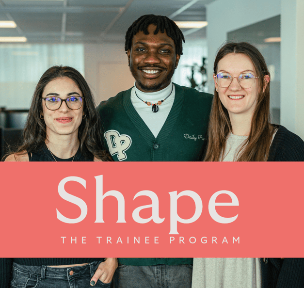 Drei lächelnde junge Kolleg:innen stehen nebeneinander in einem hellen Büro. Ein korallfarbenes Banner über dem Bild trägt die Aufschrift „Shape – The Trainee Program“.