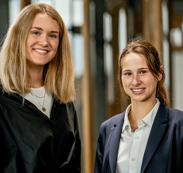 Zwei lächelnde junge Kolleg:innen stehen nebeneinander in einem modernen Büro mit Holzrahmen und Glaswänden – eine Person trägt eine schwarze Bluse, die andere einen dunkelblauen Blazer über einem weißen Hemd.