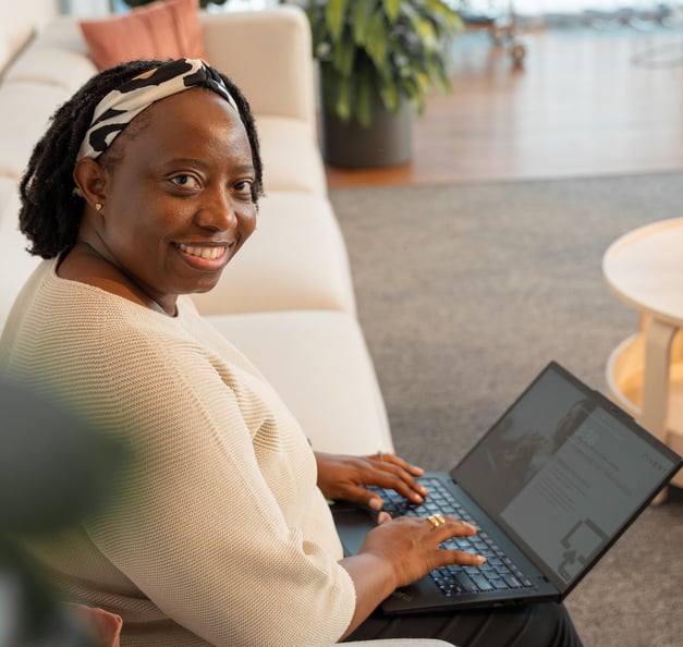 Eine Person arbeitet auf einem Sofa in einer hellen Bürolounge am Laptop und blickt in die Kamera.